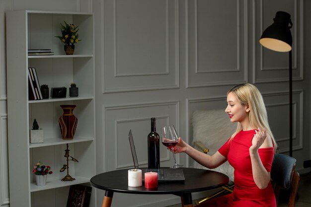 Virtuelle Liebe, süßes blondes Mädchen in rotem Kleid bei Ferndate mit Kerzen, die Wein trinken