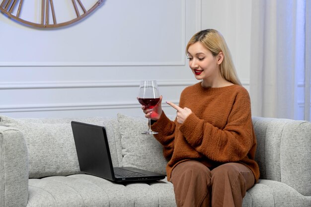 Virtuelle Liebe, süßes junges blondes Mädchen im kuscheligen Pullover auf Distanzcomputerdatum, das auf Wein zeigt