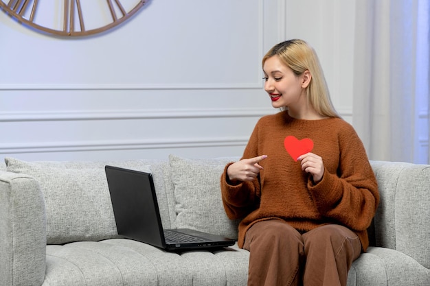 Virtuelle Liebe süßes junges blondes Mädchen im kuscheligen Pullover auf Ferncomputerdatum, das auf Herz zeigt