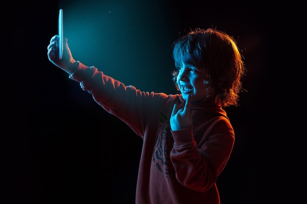 Vlogging mit Smartphone. Porträt des kaukasischen Jungen auf dunkler Wand im Neonlicht. Schönes lockiges Modell. Konzept der menschlichen Emotionen, Gesichtsausdruck, Verkauf, Werbung, moderne Technologie, Gadgets.