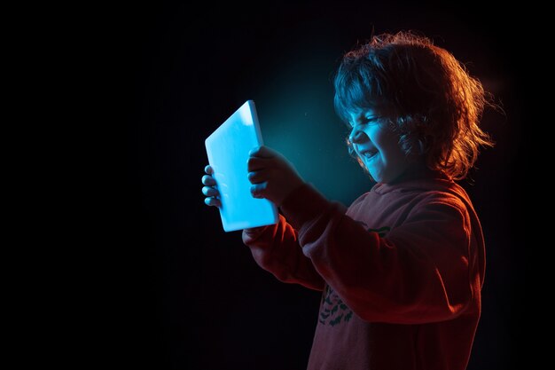 Vlogging mit Tablet, spielen. Porträt des kaukasischen Jungen auf dunkler Wand im Neonlicht. Schönes lockiges Modell. Konzept der menschlichen Emotionen, Gesichtsausdruck, Verkauf, Werbung, moderne Technologie, Gadgets.
