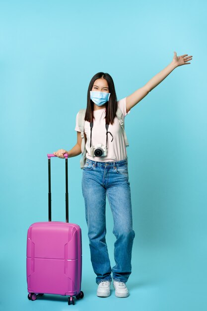Volle Länge der glücklichen koreanischen Frau in Gesichtsmaske, die mit Koffer steht, die Hand verträumt hebt, den Urlaub im Ausland genießt, auf blauem Hintergrund steht