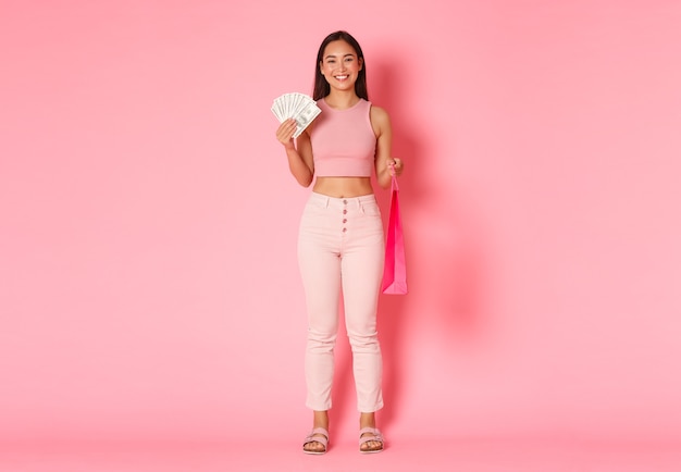 Volle Länge des attraktiven lächelnden asiatischen Mädchens in der Sommerkleidung, das Geld und die Einkaufstasche hält, etwas teures kauft, genießt, in Einkaufszentren zu gehen, rosa Wand stehend