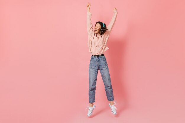 Volle Länge des Schusses der jungen Frau springend mit ihren Armen, die gegen rosa Hintergrund erhoben werden. Dame in Kopfhörern posiert und lacht.