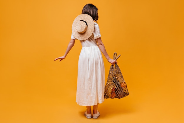 Volle Länge des Schusses des Mädchens im Midikleid mit Strohhut und Schnurbeutel mit Früchten auf orangefarbenem Hintergrund.