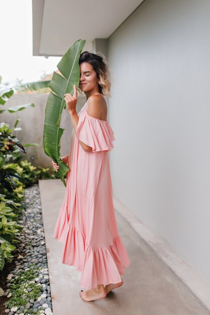 Volle Länge Schuss des anmutigen kaukasischen Mädchens, das mit grüner Pflanze aufwirft. Außenfoto der niedlichen blonden Dame trägt langes rosa Kleid.