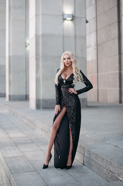 Voller Länge einer wunderschönen schlanken blonden Frau mit langen, gewellten Haaren und großer Brust, die in einem funkelnden schwarzen Kleid und High Heels auf der Straße posiert.