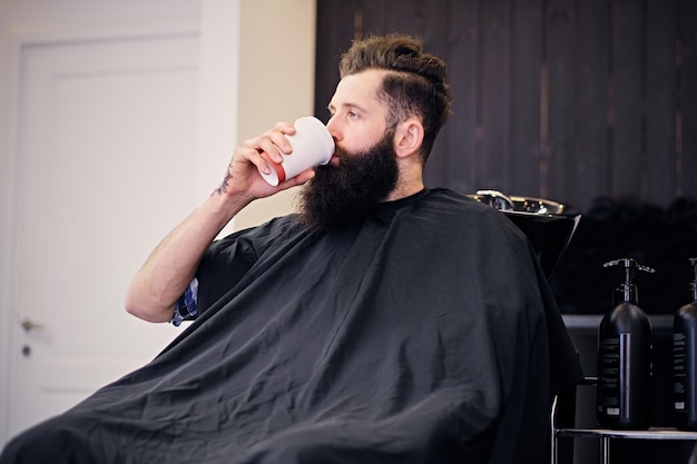 Vollgas bärtiger Hipster-Mann trinkt Kaffee vor dem Haarstyling in einem Friseursalon.
