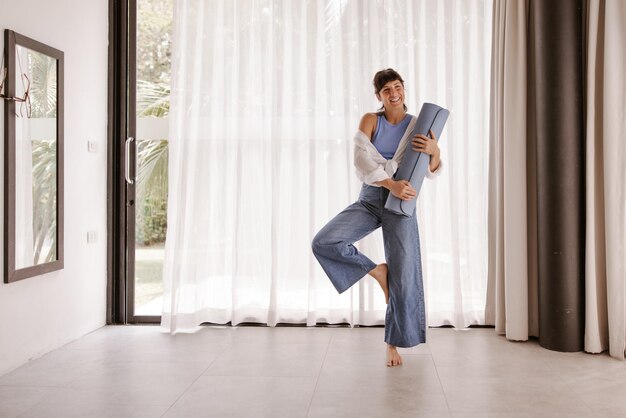 Vollständige Ansicht einer lächelnden Frau mit Fitnessmatte, die eine Sportpose macht