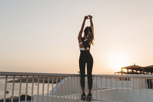 Von hinten sexy junge Frau in Sportbekleidung am Meer bei Sonnenaufgang. Sommermorgen im tropischen Land, Training, modisches Modell, Fitness, Glück.