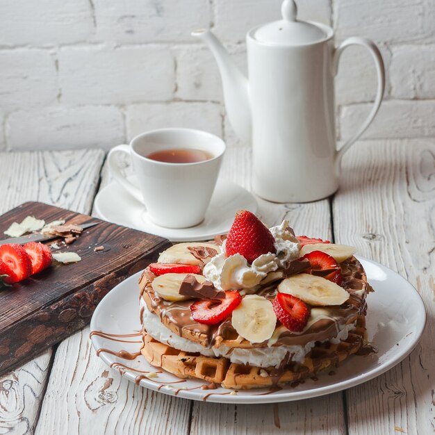 Von oben Waffeleiskuchen mit Erdbeere und Tasse und Teekanne in rundem weißen Teller