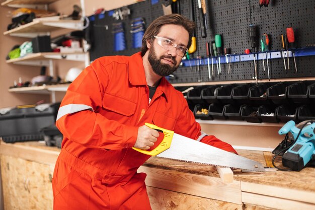 Vorarbeiter in orangefarbener Arbeitskleidung und Schutzbrille schaut nachdenklich mit der Handsäge in der Werkstatt in die Kamera