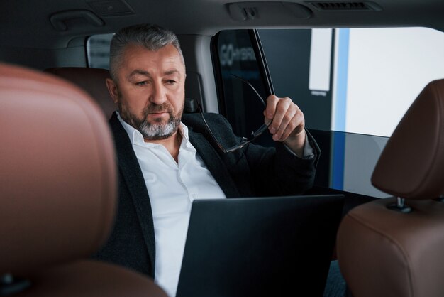 Vorbereitung zum Lesen einiger Dokumente. Arbeiten auf einer Rückseite des Autos mit silberfarbenem Laptop. Leitender Geschäftsmann