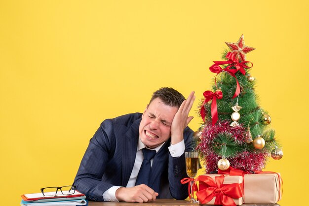 Vorderansicht aufgeregter Mann mit geschlossenen Augen, die am Tisch nahe Weihnachtsbaum und Geschenken auf gelbem Hintergrund sitzen