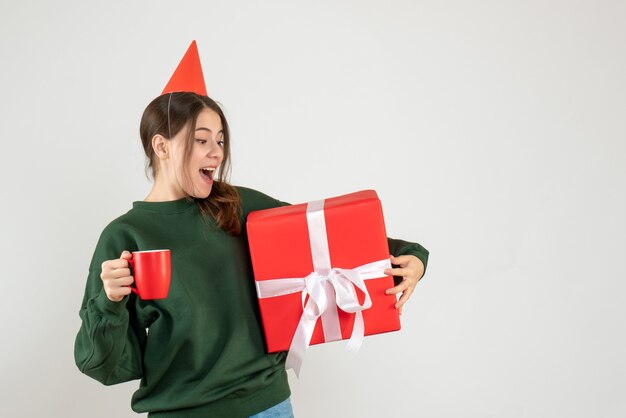 Vorderansicht aufgeregtes Mädchen mit Partykappe, die ihr Weihnachtsgeschenk und eine Tasse hält