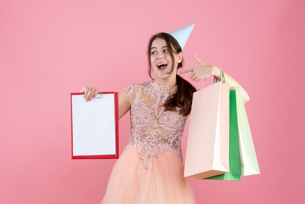 Vorderansicht begeistertes Mädchen mit Partykappe, die Dokumente und Einkaufstaschen hält, die auf sich selbst zeigen