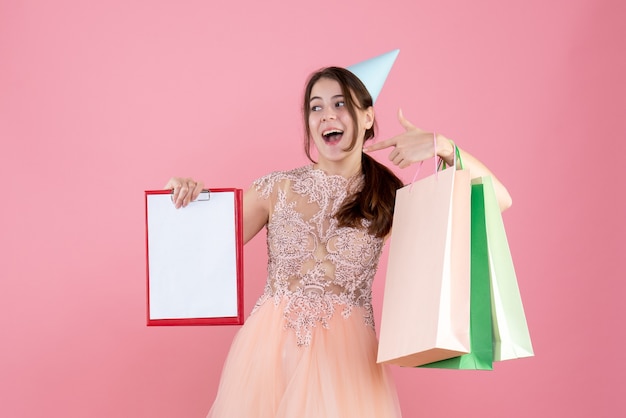 Vorderansicht begeistertes Mädchen mit Partykappe, die Dokumente und Einkaufstaschen hält, die auf sich selbst zeigen