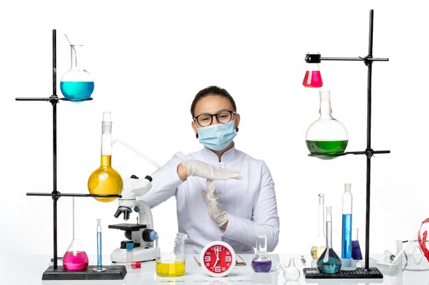 Vorderansicht Chemikerin im weißen medizinischen Anzug mit Maske sitzend mit Lösungen auf hellweißem Hintergrund Chemiker-Virus-Covid-Splash-Labor