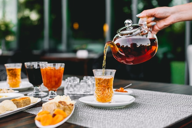 Vorderansicht das Mädchen gießt Tee aus der Teekanne in ein armoud Glas mit Marmelade und Süßigkeiten auf dem Tisch