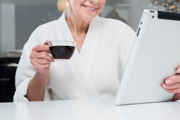 Vorderansicht der älteren Frau im Bademantel, der Tablette und Kaffeetasse hält