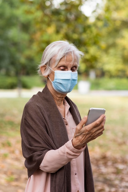 Vorderansicht der älteren Frau mit der medizinischen Maske, die Smartphone hält