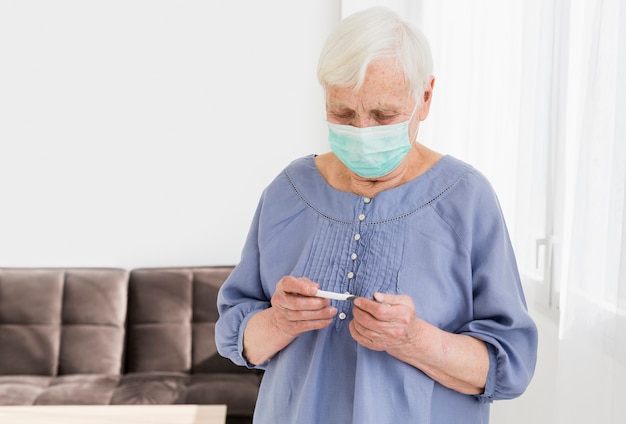 Vorderansicht der älteren Frau mit der medizinischen Maske, die Thermometer betrachtet