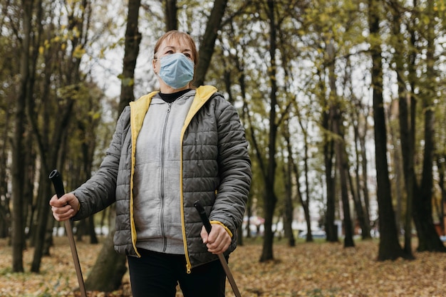 Vorderansicht der älteren Frau mit medizinischer Maske und Trekkingstöcken im Freien