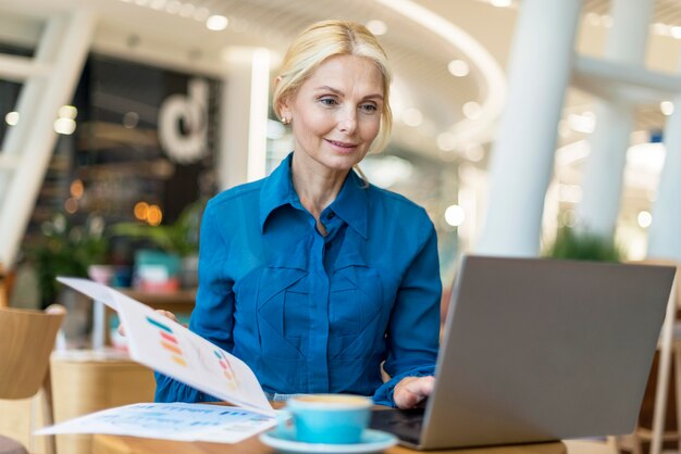 Vorderansicht der älteren Geschäftsfrau, die am Laptop mit Kaffee und Papieren arbeitet