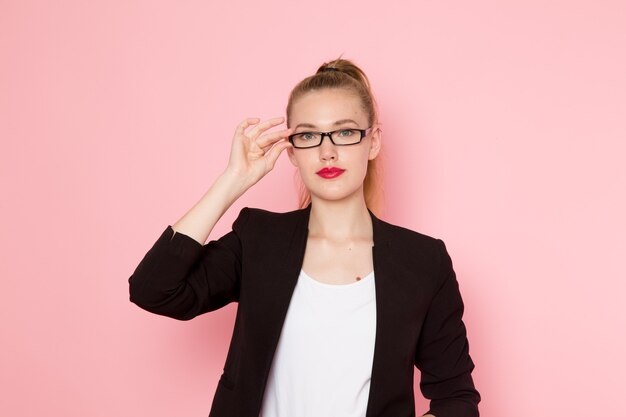 Vorderansicht der Büroangestellten in schwarzer strenger Jacke, die ihre Sonnenbrille auf hellrosa Wand berührend aufwirft