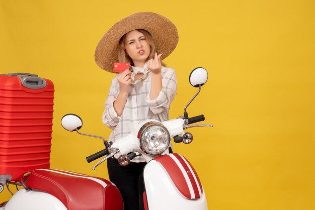 Vorderansicht der ehrgeizigen jungen Frau, die Hut trägt, der ihr Gepäck sammelt, das auf Motorrad sitzt und Bankkarte hält, die Geldgeste macht