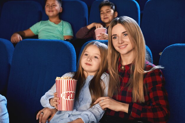 Vorderansicht der Familie, die Zeit zusammen im Kino verbringt. Attraktive junge Mutter und kleine Tochter, die sich umarmen und lächeln, während sie Film schauen und Popcorn essen