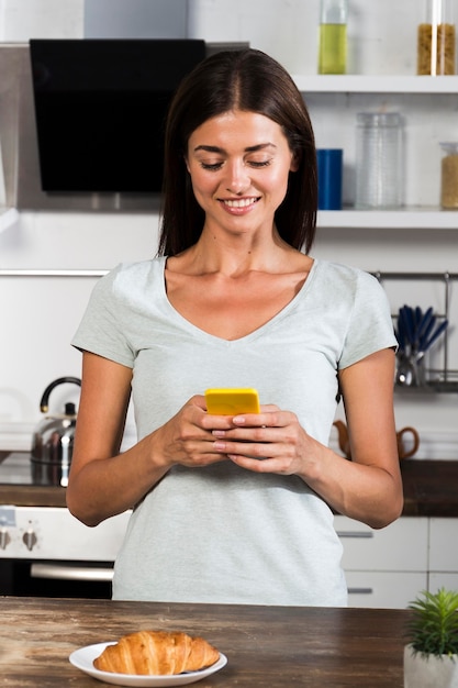 Vorderansicht der Frau in der Küche mit Smartphone und Croissant