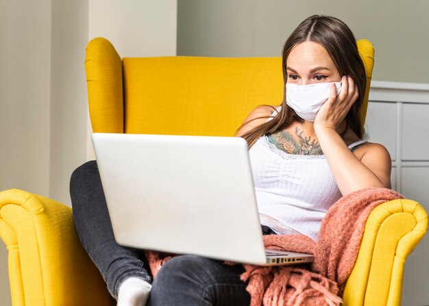 Vorderansicht der Frau mit der medizinischen Maske, die am Laptop vom Sessel während der Pandemie arbeitet