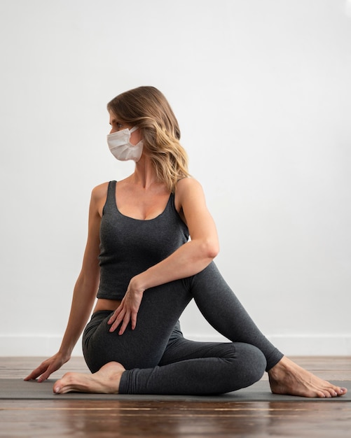Vorderansicht der Frau mit der medizinischen Maske, die Yoga zu Hause tut