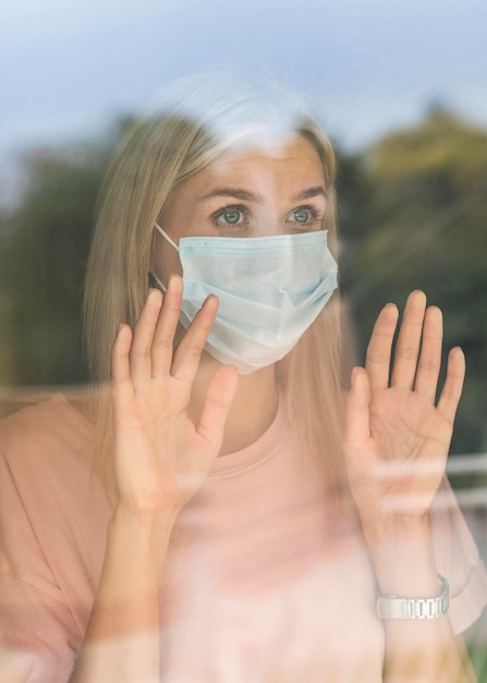 Vorderansicht der Frau mit der medizinischen Maske zu Hause, die Fenster während der Pandemie berührt