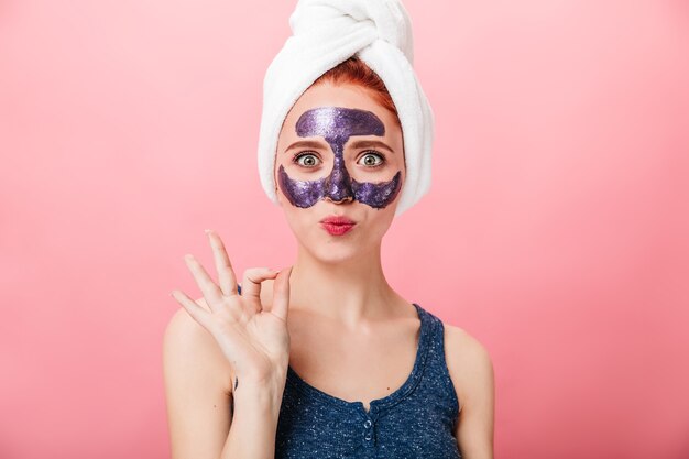 Vorderansicht der Frau mit Gesichtsmaske, die okay Zeichen zeigt. Studioaufnahme des verblüfften Mädchens mit Handtuch auf Kopf, das auf rosa Hintergrund gestikuliert.