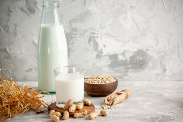 Vorderansicht der Glasflasche und Tasse gefüllt mit Milch auf Holztablett und Trockenfrüchtelöffel Hafer in braunem Topf auf der linken Seite auf weißem Tisch auf Eishintergrund