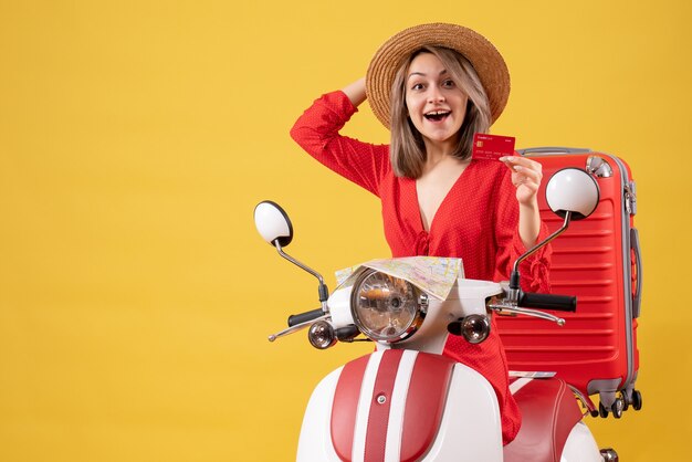 Vorderansicht der glücklichen jungen Dame im roten Kleid, die Bankkarte auf Moped hält