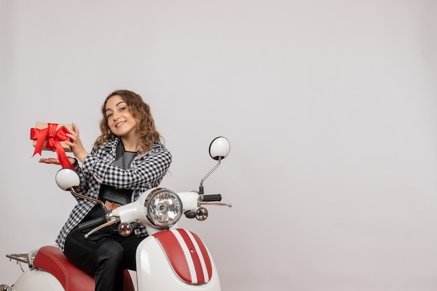 Vorderansicht der glücklichen jungen Frau auf Moped, das Geschenk auf grauer Wand hält
