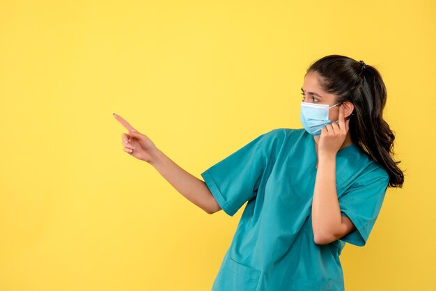 Vorderansicht der hübschen Ärztin mit der medizinischen Maske, die links auf gelbe Wand zeigt