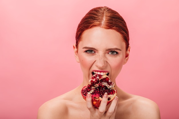 Vorderansicht der hungrigen frau, die granatapfel isst. studioaufnahme des nackten ingwermädchens mit granat lokalisiert auf rosa hintergrund.