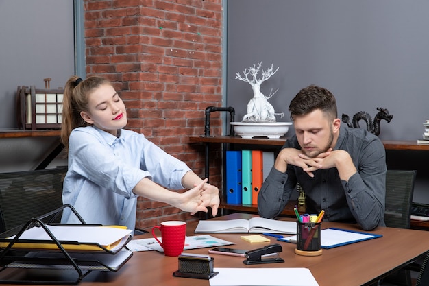 Vorderansicht der jungen Arbeiterin und ihres männlichen Kollegen, der müde am Tisch im Büro sitzt sitting