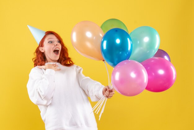 Vorderansicht der jungen Frau, die bunte Luftballons an der gelben Wand hält