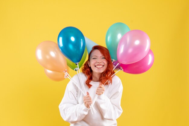 Vorderansicht der jungen Frau, die bunte Luftballons an der gelben Wand hält