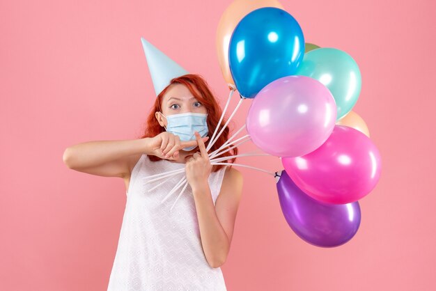 Vorderansicht der jungen Frau, die bunte Luftballons in der sterilen Maske auf rosa Wand hält