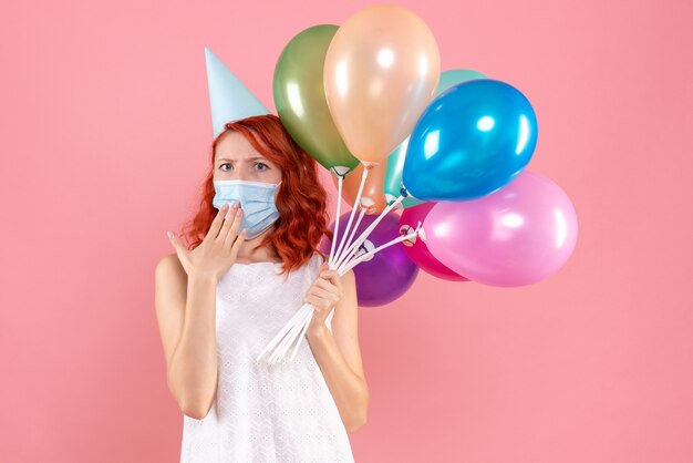 Vorderansicht der jungen Frau, die bunte Luftballons in der sterilen Maske auf rosa Wand hält