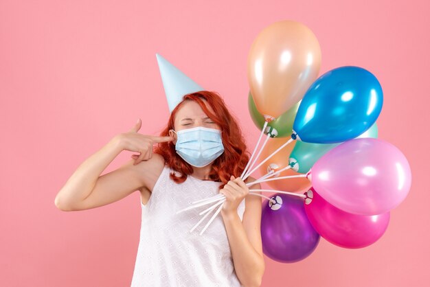 Vorderansicht der jungen Frau, die bunte Luftballons in der sterilen Maske auf rosa Wand hält