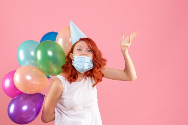 Vorderansicht der jungen Frau, die bunte Luftballons in der sterilen Maske auf rosa Wand hält