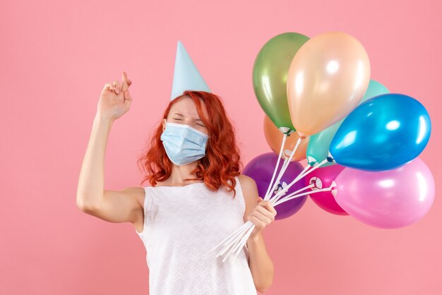 Vorderansicht der jungen Frau, die bunte Luftballons in der sterilen Maske auf rosa Wand hält