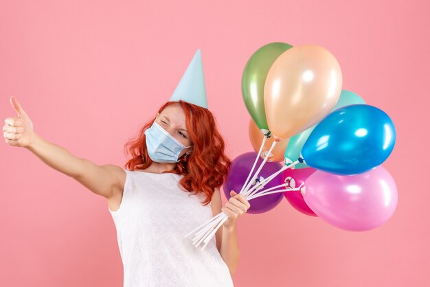 Vorderansicht der jungen Frau, die bunte Luftballons in der sterilen Maske auf rosa Wand hält